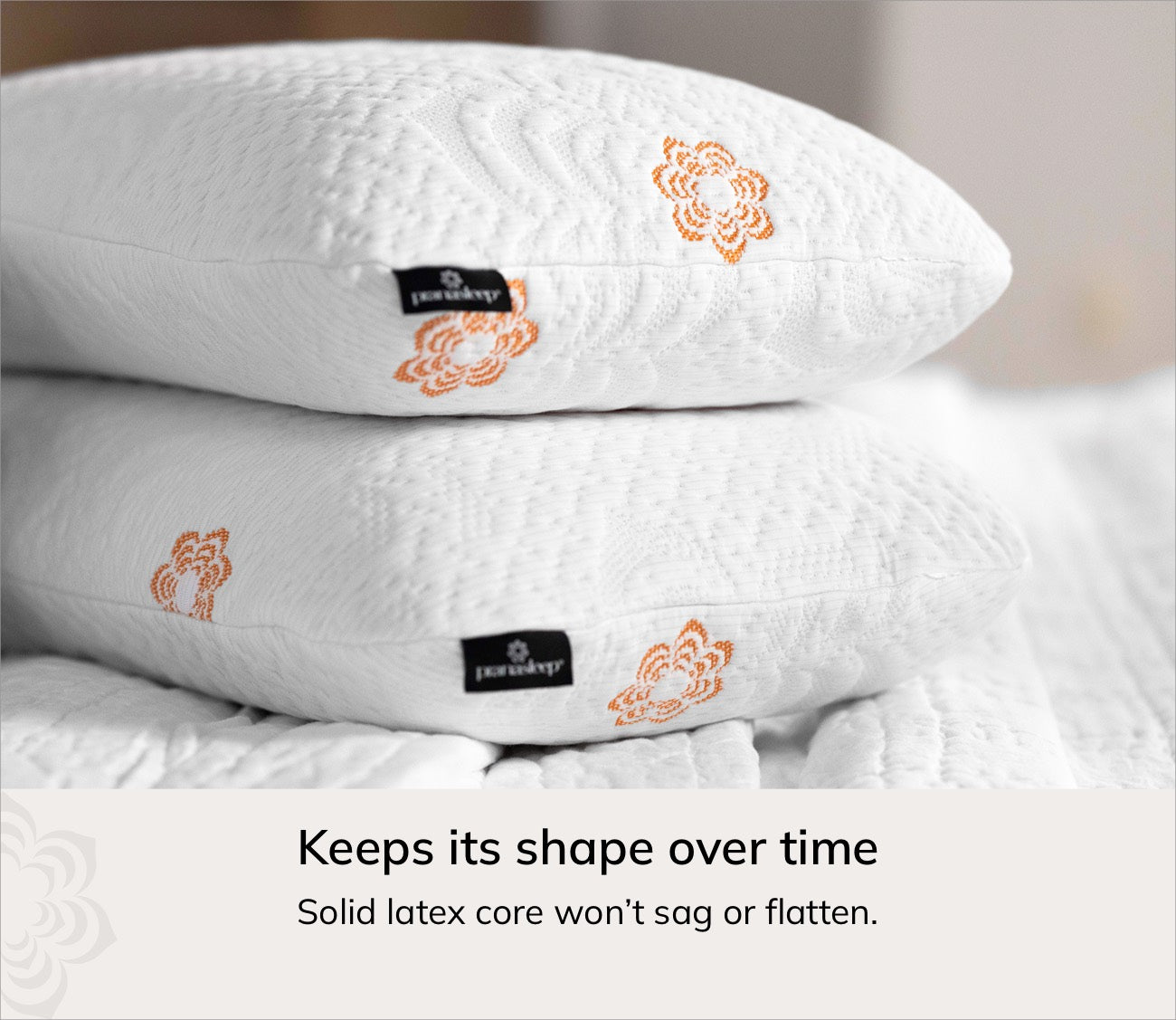 Two stacked karma pillows with orange floral embroidery and black brand tags reading 'KESPERA' on a soft bed background, with text below stating it keeps its shape over time.