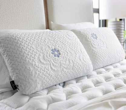 Two PranaSleep Asha Natural Latex Pillows displayed on a bed, showcasing their textured knit covers and signature blue embroidered details.